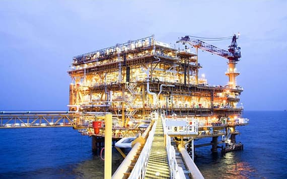 Lit offshore oil and gas platform at dusk, jetty extending across calm water — the kind of capital-heavy infrastructure that's failing without an intelligence layer.